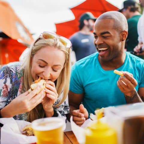 A couple laughing while eating burgers and fries and drinking beer at an outdoor restaurant